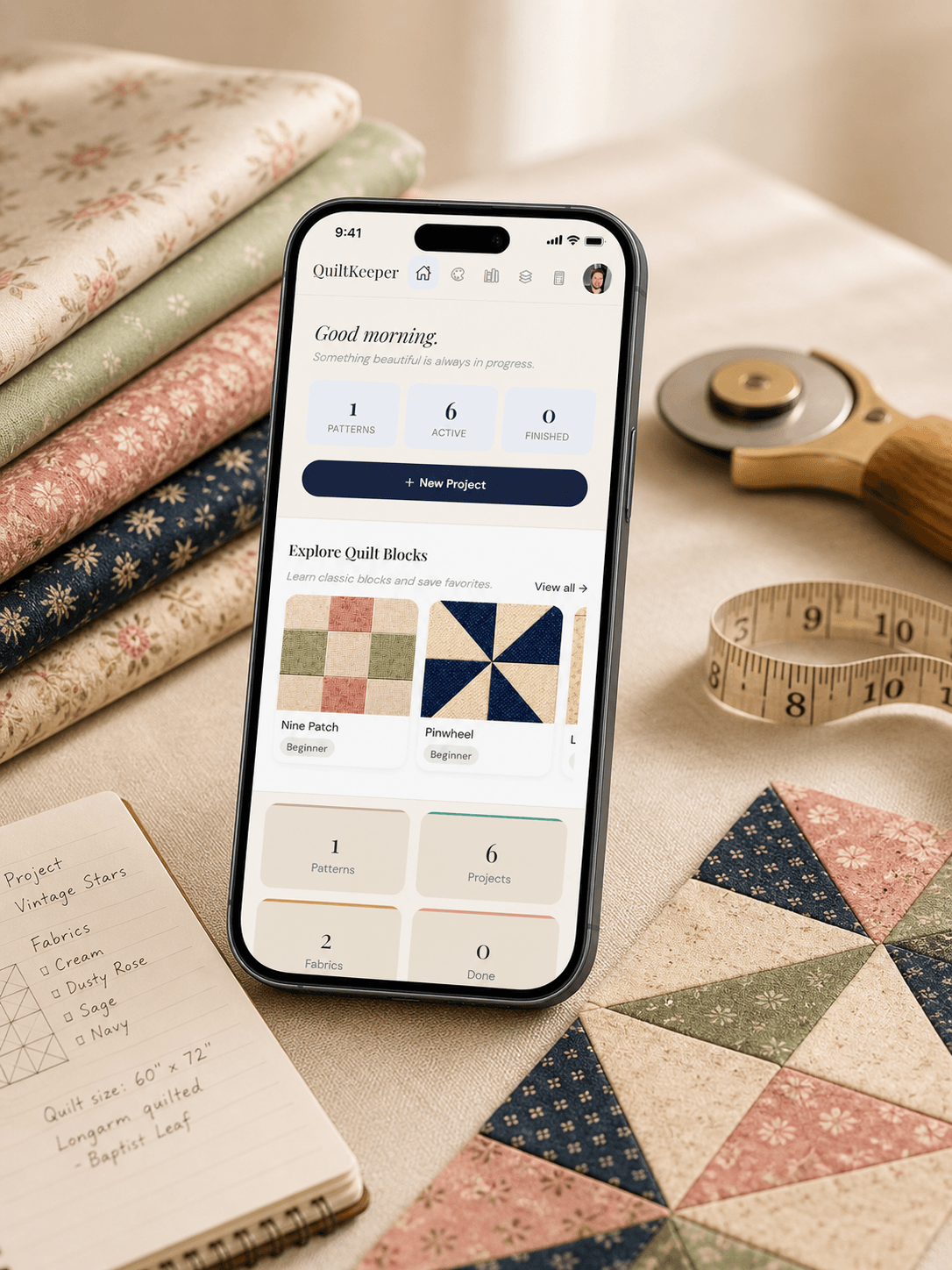 QuiltKeeper app open on an iPhone, surrounded by folded fabric swatches and an open project notebook on a warm cream surface